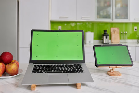 Two wireless mobile gadgets with black crosses on green screens standing on wooden holders on white marble kitchen tableの写真素材