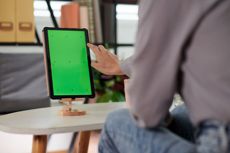 Hand of young woman going to touch blank green screen of tablet on wooden holder while sitting in front of mobile gadgetの写真素材