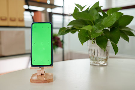 Smartphone with blank green screen standing on wooden holder on white table against bunch of leaves of domestic plantの写真素材