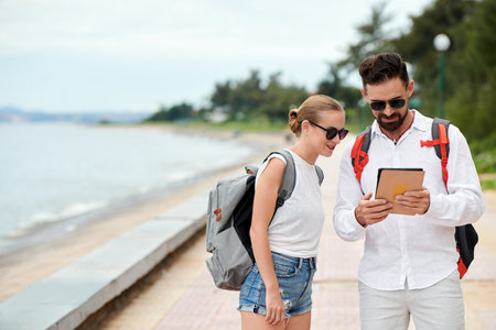Young couple of tourists with backpacks using digital tablet while standing near the coastline outdoorsの写真素材