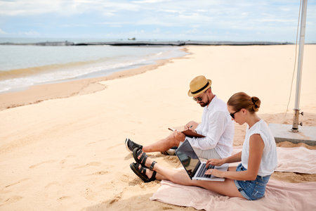 Young couple sitting on the beach together and working online using laptopの写真素材