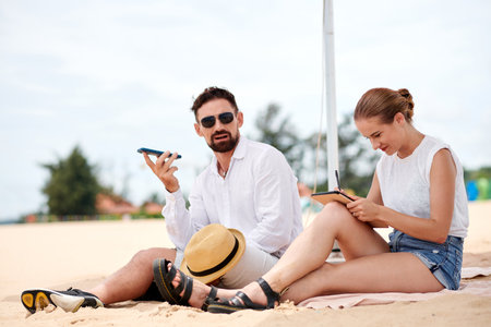 Young couple working together while sitting on the beach, they using mobile phone and tablet pcの写真素材