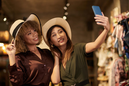 Happy friends wearing hats posing at camera of mobile phone while doing shopping in mallの写真素材