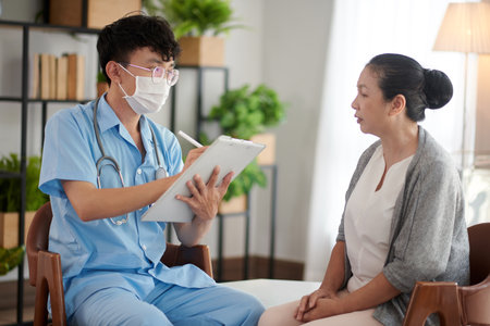 Doctor in medical mask talking to patient of nursing home and taking notes in documentの写真素材