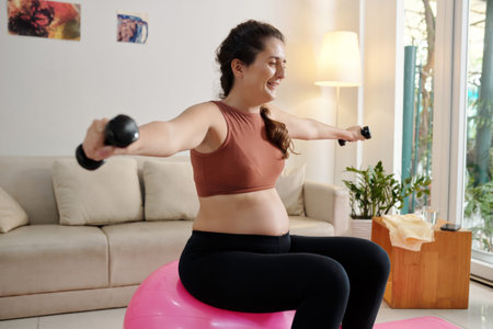 Cheerful pregnant woman sitting on fitness ball and doing exercise with small dumbbells