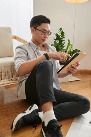 Smiling young architect sitting on floor and reading article on tablet computer