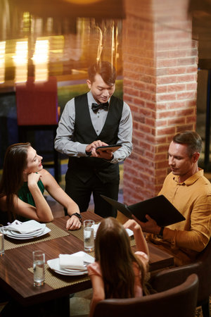 Smiling waiter taking notes on tablet computer when family ordering food