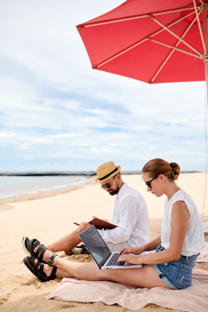 Digital nomads sitting under beach umbrella, working on laptop and reading bookの写真素材