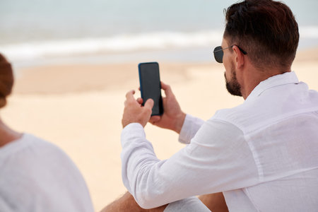 Businessman in white shirt sitting on beach and checking messages and notifications on smartphoneの写真素材