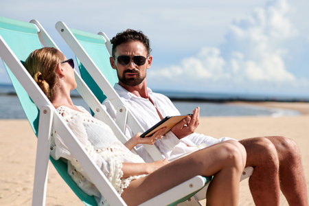 Frowning man talking to girlfriend when they are sunbathing in chaise-lounges on beach