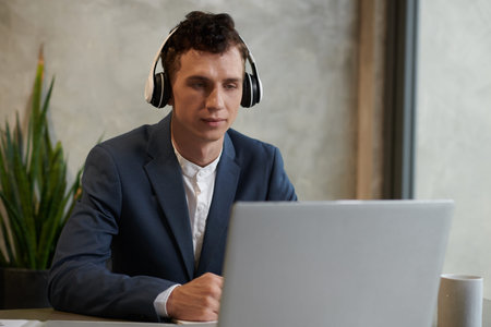 Smiling young businessman listening to music in headphones when reading report on laptop screen
