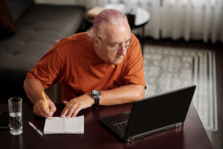 Senior man watching educational video on laptop and taking notes in notebookの写真素材