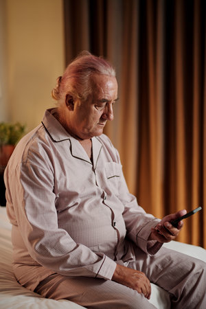 Senior man in satin pajamas sitting in bed and checking messages and notifications in smartphoneの写真素材
