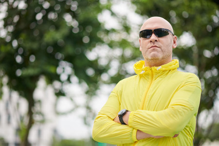 Serious mature jogger in sunglasses standing in park with arms crossed