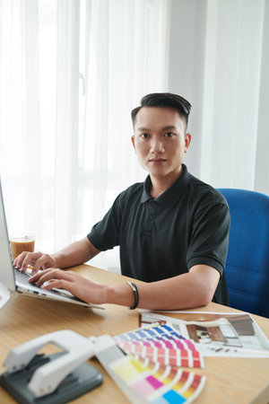 Portrait of young Vietnamese interior designer working on laptop at his office tableの写真素材