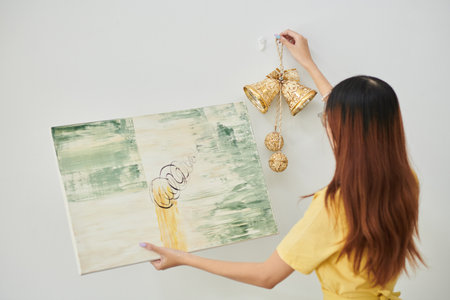 Young woman hanging golden bells and abstract picture on wall in apartment, view from backの写真素材