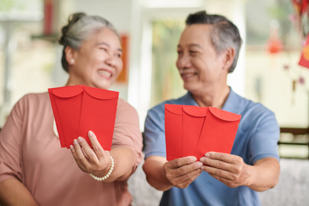 Positive senior man and woman showing red lucky money envelopes and looking at each otherの写真素材