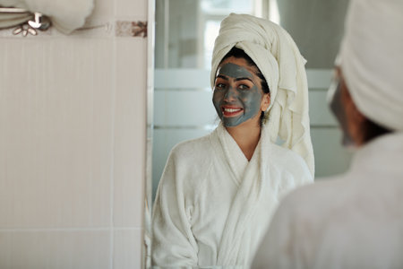 Cheerful young woman with charcoal mask on face looking at bathroom mirrorの写真素材