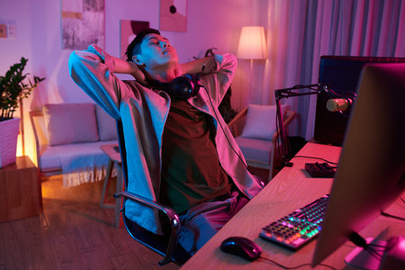 Tired teenage boy stretching back and leaning back in his chair after finishing raid in videogame