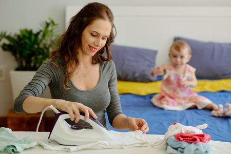 Smiling young woman ironng baby clothes when daughter playing in backgroundの写真素材