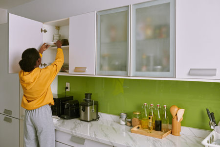 Woman in homewear taking plate out of kitchen cabinet, view from the back