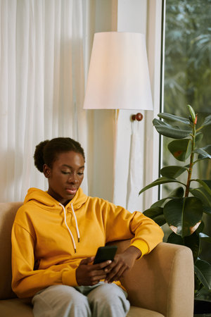 Black young woman sitting on couch and answering text messages from friends and family membersの写真素材