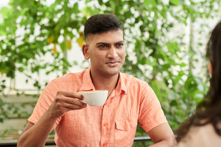 Portrait of Indian man drinking cup of cappuccino when meeting with friend in outdoor cafeの写真素材