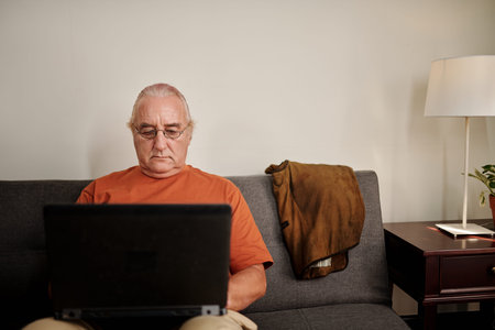 Portrait of elderly man in glasses sitting on couch and reading document on laptop screen