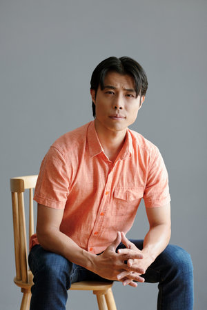 Confident young Asian man sitting on chair against grey wallの写真素材