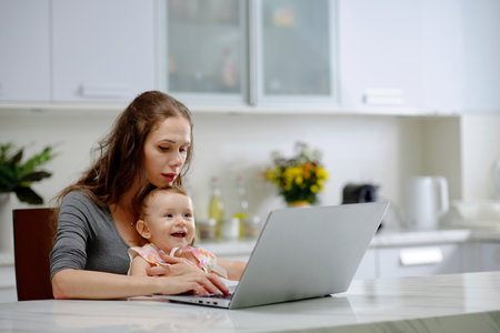 Young woman working on laptop with little daughter sitting on her lapsの写真素材