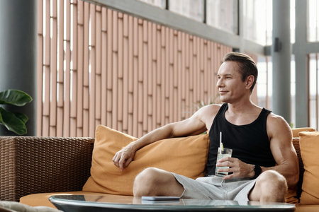 Smiling sportsman resting on couch and drinking ice water after trainingの写真素材