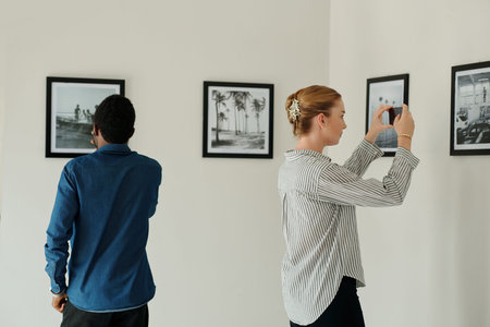 Young woman with smartphone taking photo of picture on wall while standing in art gallery against African American male visitorの写真素材