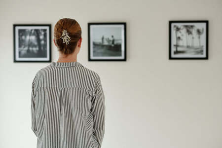 Rear view of young woman in striped shirt standing in front of wall with pictures or artworks in black frames while visiting art galleryの写真素材
