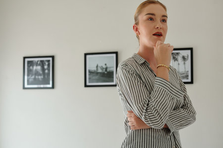 Young woman in casualwear looking at artwork with attention while standing in front of camera against wall with row of picturesの写真素材