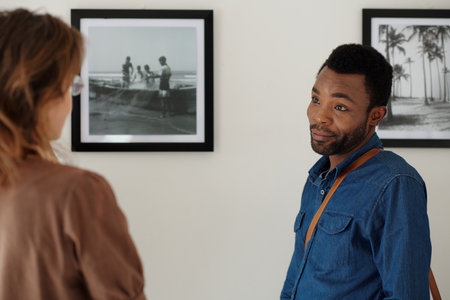 Young African American male guest of modern art gallery talking to woman while standing by wall with two black-and-white picturesの写真素材