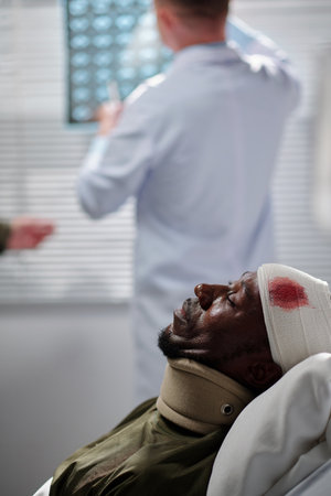 Wounded African American patient with injured head lying on bed in ward with doctor examining x-ray image in backgroundの写真素材