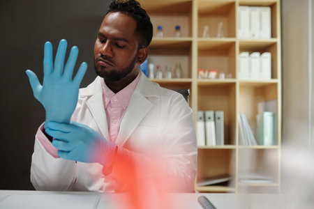 Young male scientist in lab coat sitting by workplace in laboratory and putting on protective gloves before working with liquidsの写真素材