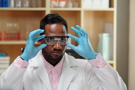 Young black man in lab coat and gloves putting on protective eyeglasses before carrying out...