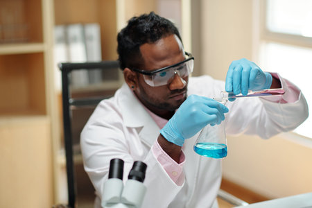 Young black man in workwear pouring liquid from flask into beaker while sitting by workplace in...