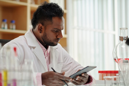 Young serious male scientist with tablet looking at blood samples in flasks while sitting by workplace in laboratory and studying new virusの写真素材