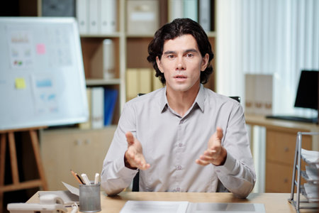 Young confident male business coach explaining something at seminar while sitting by desk in front...