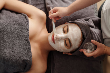 Cosmetologist applying brightening white clay mask on face of young womanの写真素材