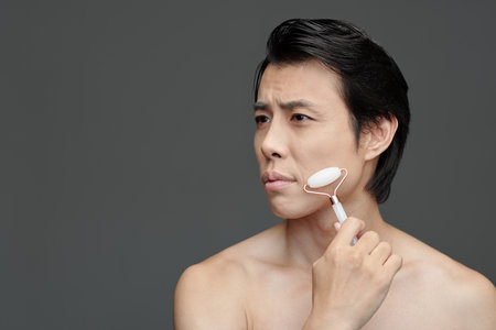 Frowning Asian man massaging face with roze quartz rollerの写真素材