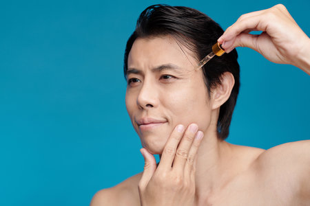 Man applying vitamin c serum on face, isolated on blueの写真素材