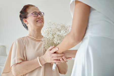 Happy senior woman touching hands of daughter standing in wedding dress ready for ceremonyの写真素材
