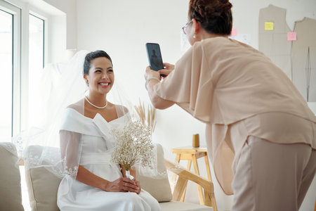 Happy bride posing for photo when mother photographing on smartphoneの写真素材
