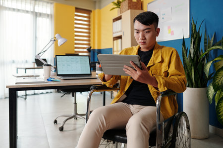 Young man with disability reading article on tablet computer when working in officeの写真素材