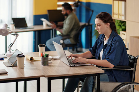 Serious young woman with disability working on laptop, answering e-mails from colleagues