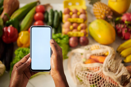 Hands holding smartpone with empty screen over counter with fresh fruits and vegetablesの写真素材