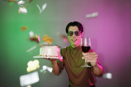 Joyful man with birthday cake and glass of wine standing under falling confettiの写真素材